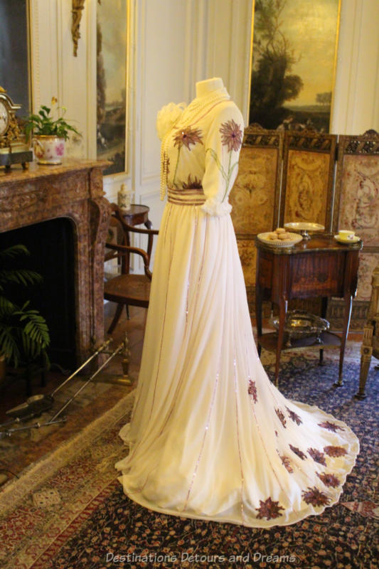 Mannequin in front of fireplace wearing long white flowing gown with embroidered flowers on bodice and train 