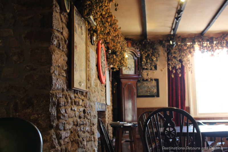 English country pub interior with one stone wall, timber beams on ceiling, hanging hops, and dark wood tables and chairs
