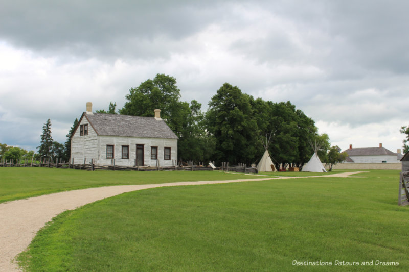 Wooden farm manager's cottage beside tepees on field outside Lower Fort Garry