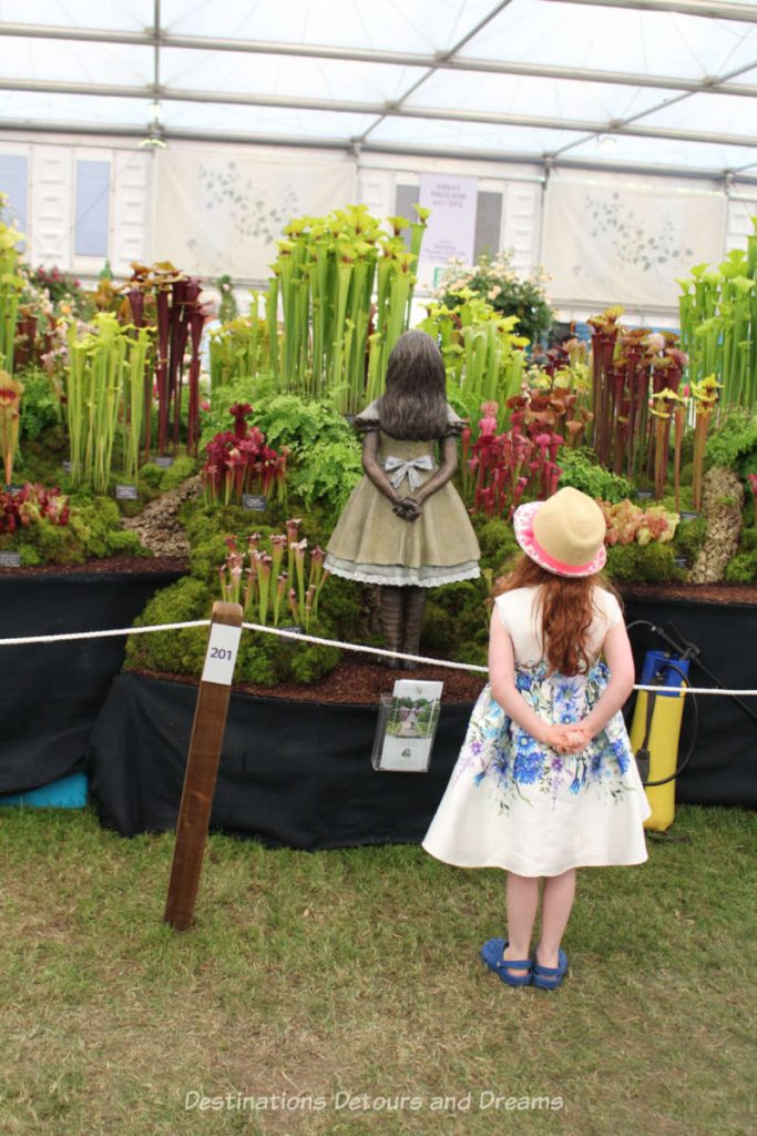 Girl watching a statue of a girl looking at a group of coniferous plants at the Chelsea Flower Show