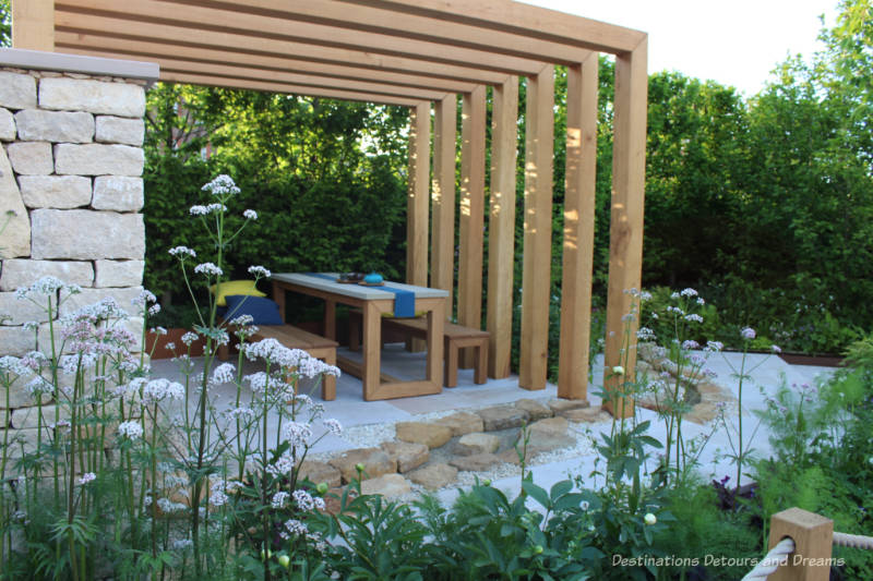 Table and beach in an outdoor seating area covered by a pergola in the Kampo no Niwa Garden at the 2019 Chelsea Flower Show