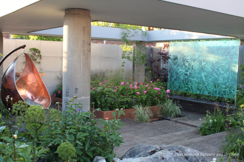 Raised garden bed in area of garden stones with a botanical water wall and bronze hanging chair in the Silten Pool Gin Garden at the Chelsea Flower Show