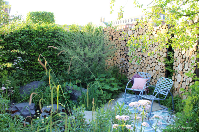 Chairs in a patio area with a wood log wall behind and a pond and wetland plants in front in The Art of Viking Garden at the 2019 Chelsea Flower Show