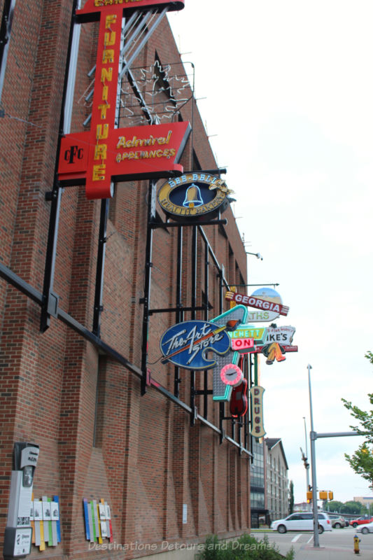 Neon signs along a brick with placards at eye level explaining their history at the Edmonton Neon Museum Signs