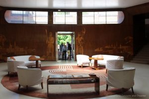 White sofas, reddish-brown circular rug with geometric design, wood wall with marquetry figures and landscapes in Eltham Palace entrance hall