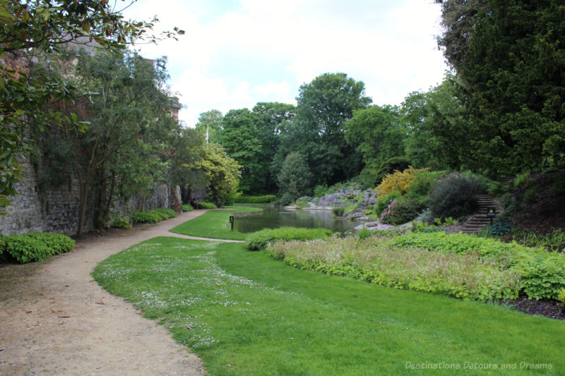 Pathway alongside brick wall beside garden green space and pond at Eltham Palace