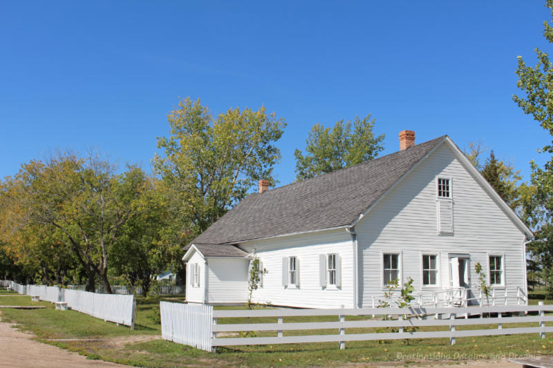 Plan white wooden church with no ornamentation in yard surrounded with a low white fence.