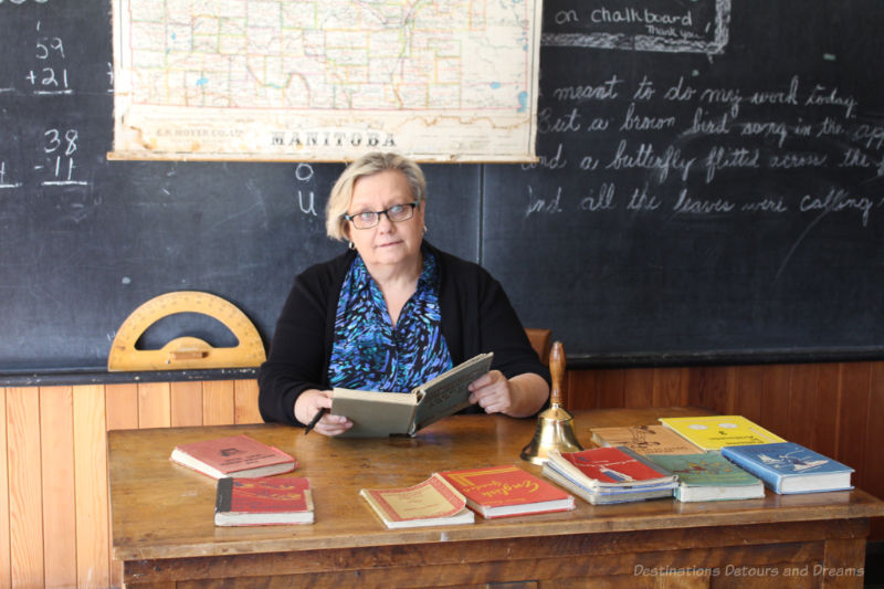 Women with book sitting behind teacher desk in a one-room school