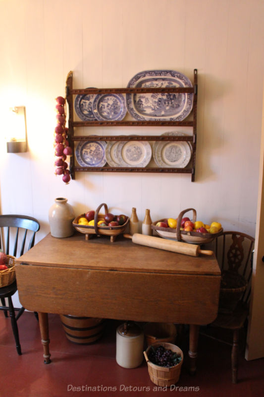 Kitchen table with baskets of fruit on it and a shelf above holding blue and white plates