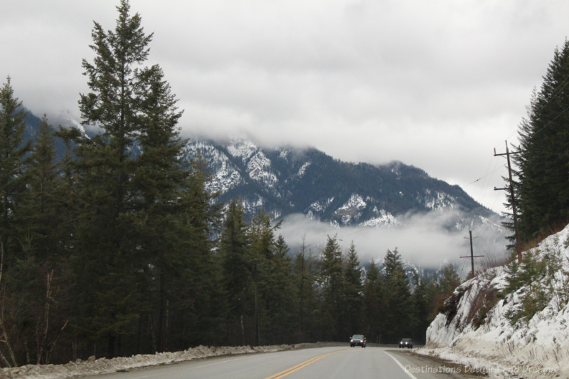 Highway lined with fir trees amid snow-covered mountains with low-lying clouds on a winter drive through British Columbia