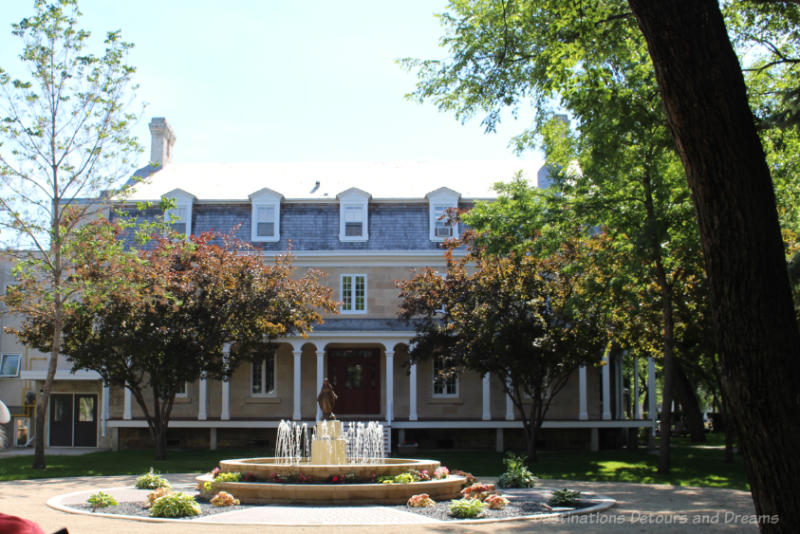 Circular drive with garden and fountain in centre in front of two-story house with wide full-width veranda and white columns