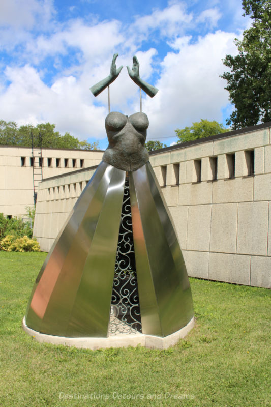 A sculpture of a dress with a wide metal skirt, a fitted bodice and disconnected hands above the dress reaching to the sky