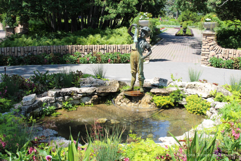 A bronze statue of a boy with a boot at the edge of a circular fountain surrounded by floral plantings