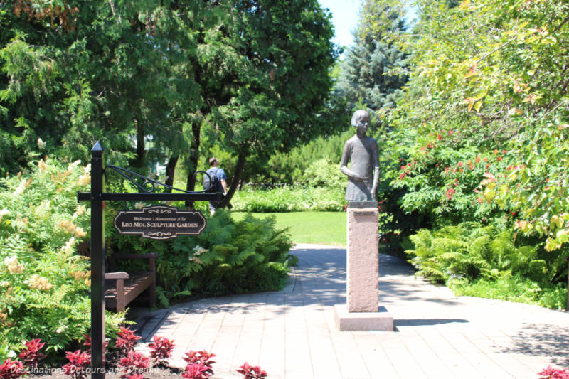 Sculpture of a school girl on a pedestal along a stone pathway surrounded by trees and shrubs at the entrance to a sculpture garden