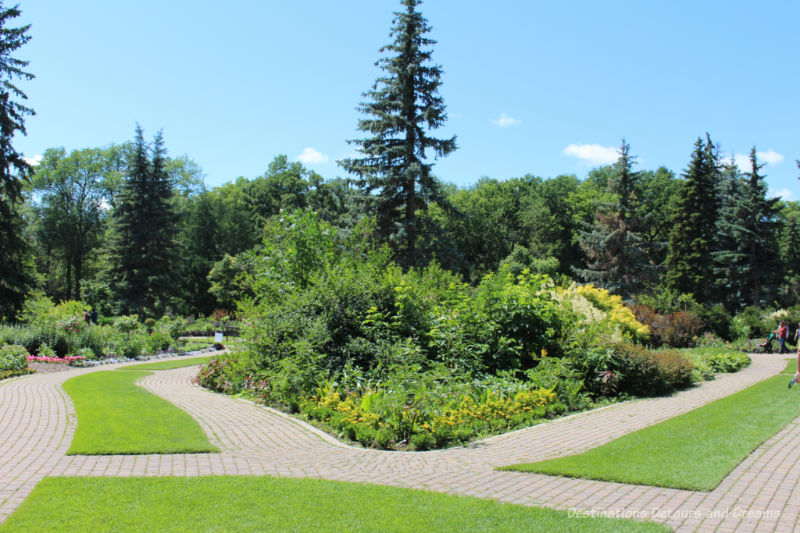 Stone double-wide pathways separated by sod patches run along either side of triangular planting of greenery and shrubs