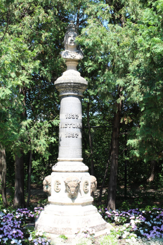 Stone pillar statue with head of Queen Victoria at the top. Statue is set in a garden and is surrounded by trees.