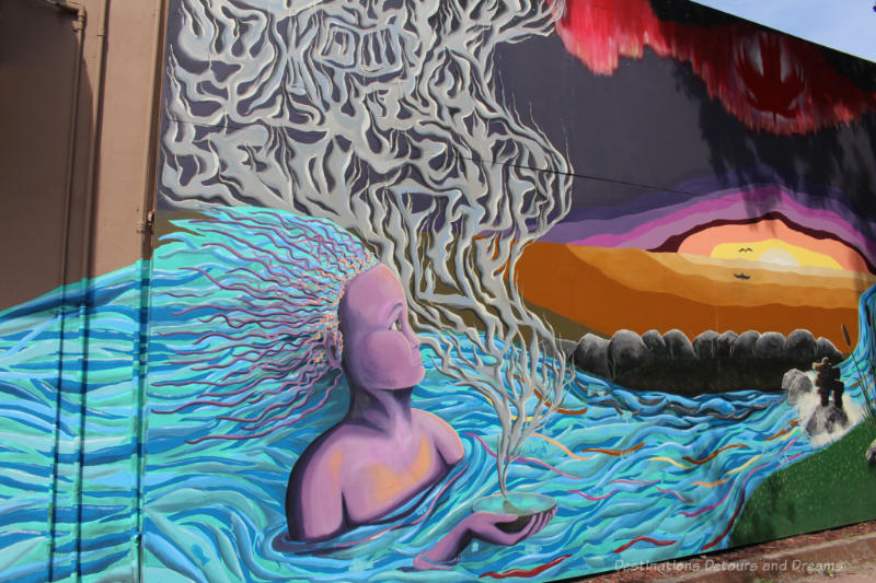 Part of a mural featuring a stylized person with flowing blue-green hair representing bodies of water doing a sage grass smudge again a background of rocks and coloured sky.