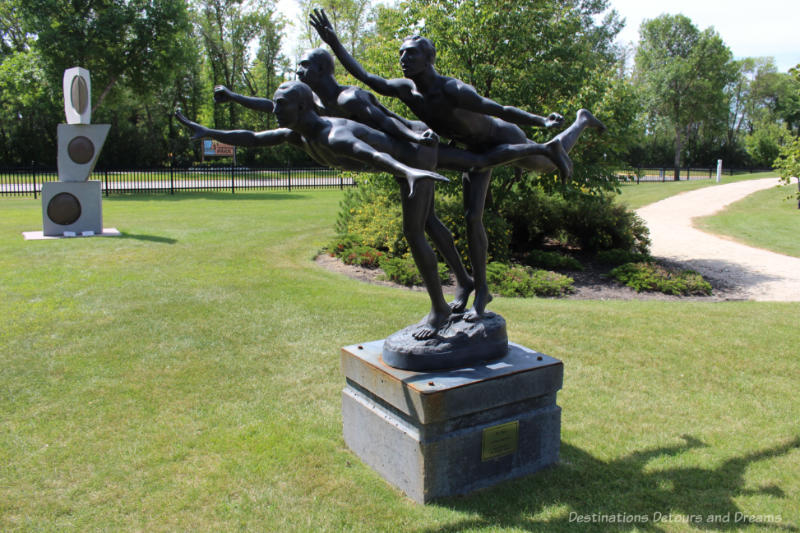 A statue in a park features three men  close together, the arms and one leg of each outstretched