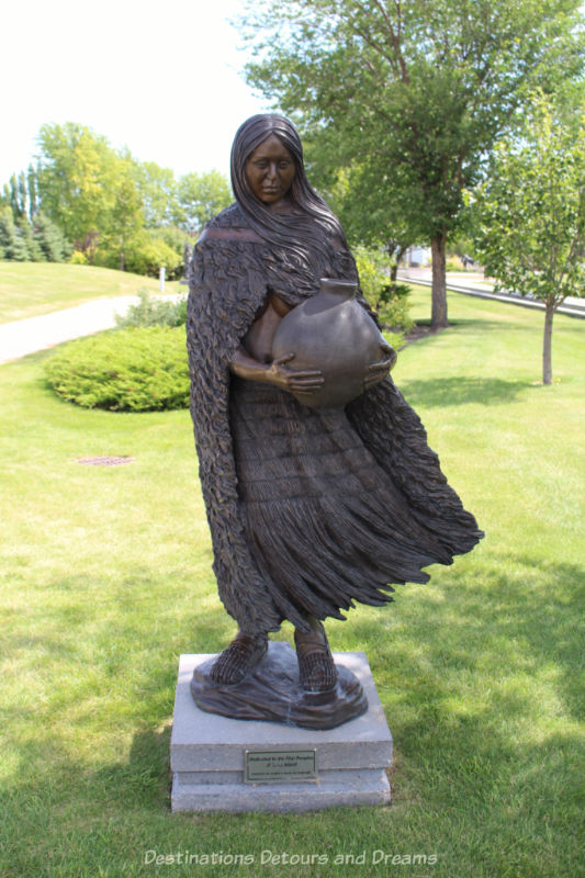 A sculpture in a park showing an Indigenous woman with flowing hair and dress holding a water jug