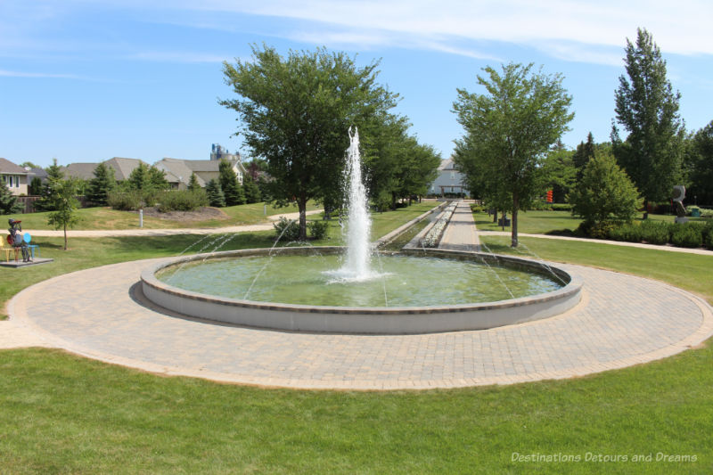 A circular fountain surrounded by a paving stone walkway with the walkway leading behind the fountain through a park to a heritage building