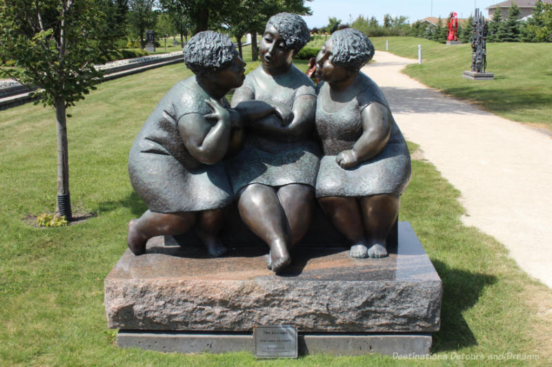 A sculpture beside a gravel walkway in a park featuring three chubby women seated and huddled together talking