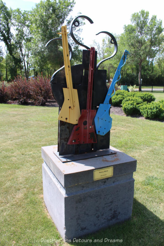 A sculpture in a park features three guitar-like instruments (yellow, red, and blue)