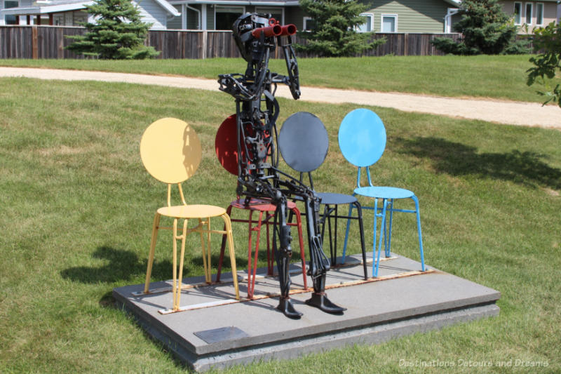 A sculpture in a park features four coloured metal chairs (yellow, red, black, blue) with round seats and backs and with a black metal skeleton sitting in the red chair looking out through red binoculars