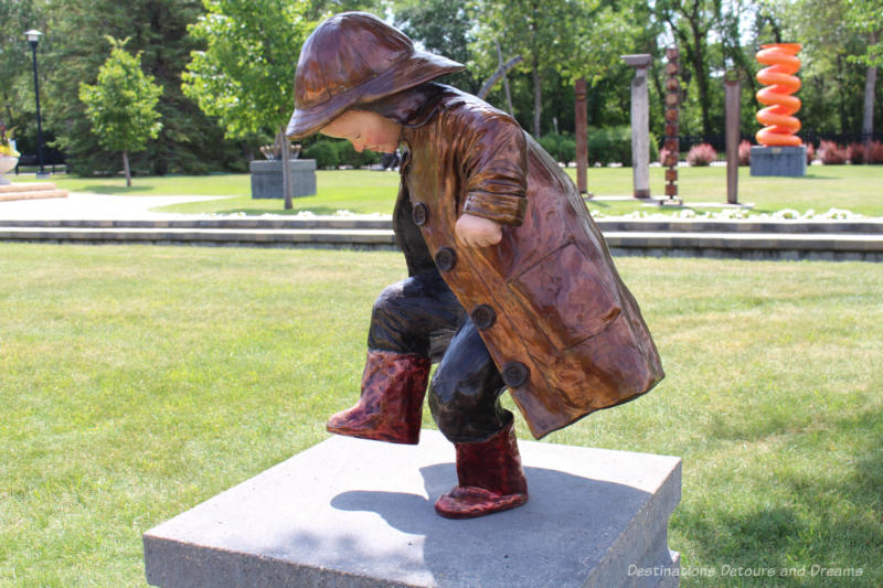 A bronze sculpture of a little boy in boots, a raincoat, and rain hat with one foot raised as if about to step in or over a puddle