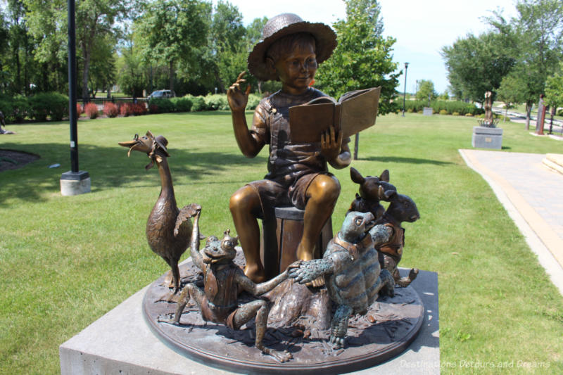 A bronze sculpture in a park features a little boy sitting and reading a book surrounded by playful animals - a goose in a bonnet, a squatting frog, a turtle in a bow tie, and more