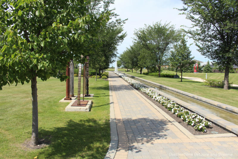 A paving stone walkway in a park containing sculptures is bordered by a flower bed and water stream, and leads to a fountain