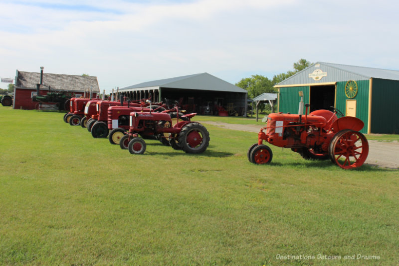 Red vintage tractors parked in a row in a field with several hangar type buildings in the background