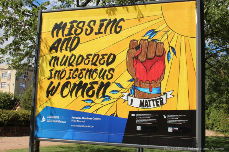 Painting with a brown coloured fist with palm covered in blood and a banner below it saying I Matte, and the words Missing and Murdered Indigenous Women printed against a background of yellow stripe emanating from a sun at the top right