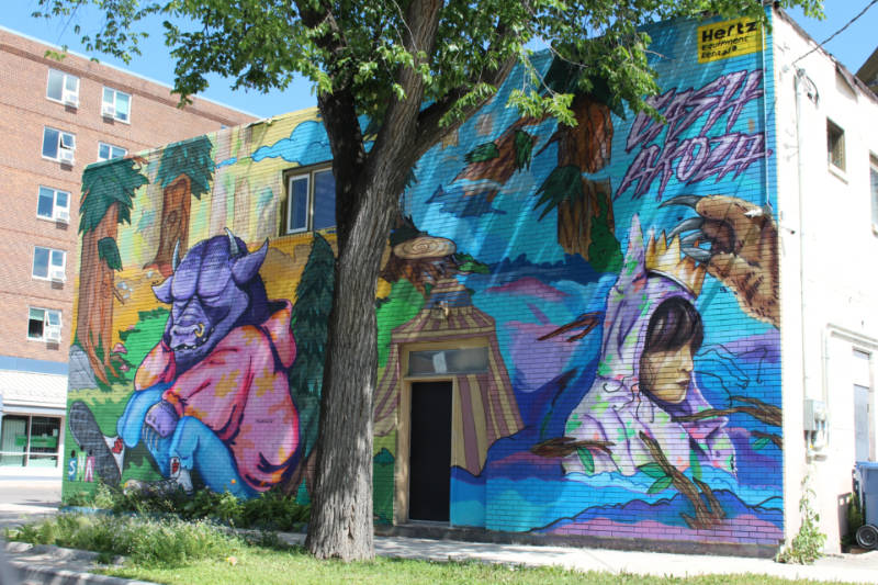 Mural in shades of blue, pink, and purple depicting a clothed animal with big ears and small horns in front of trees and woman with a colourful headscarf being crowned by a hairy hand with long talon nails