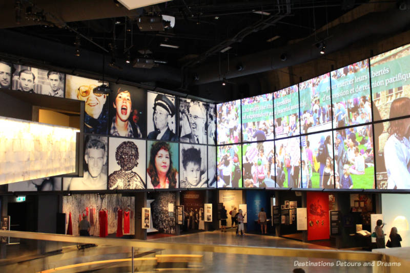 Looking down at niches of museum displays on the outside walls of a large room with images of people and human rights symbols projected on the tall walls above them at the Canadian Museum for Human Rights