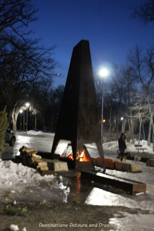 Fire blazing at the bottom of a tall steel fire pit public art piece on a winter night