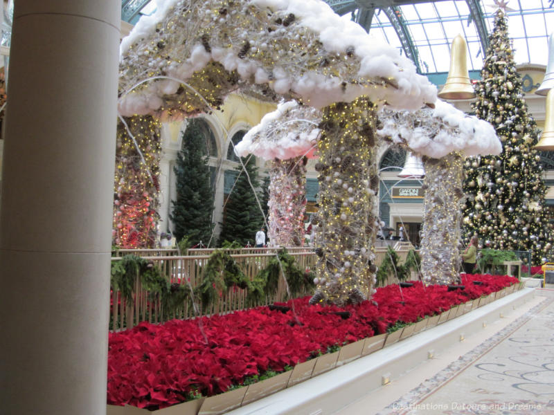 A bed of red poinsettias, columns covered in gold and white balls and lights holding up a roof of the same balls and lights trimmed with white to look like snow in a Christmas display in Bellagio Hotel