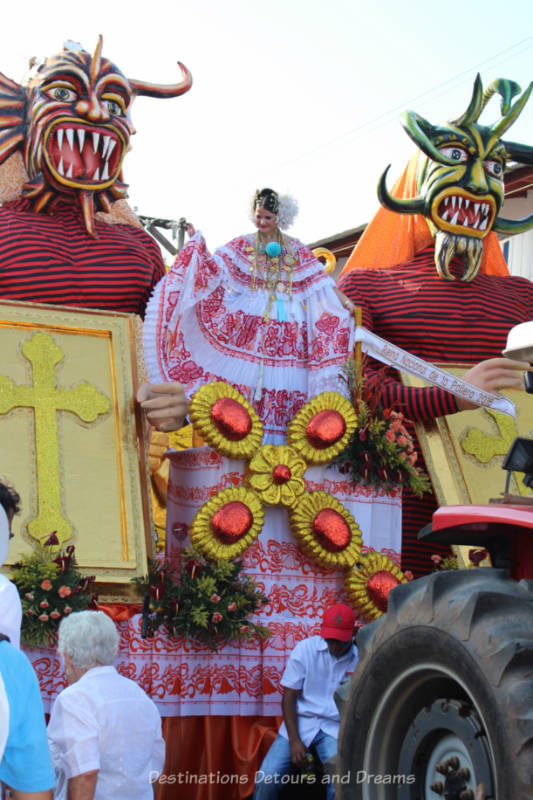 Parade float featuring devil mask characters flanking a woman in a red and white pollera dress