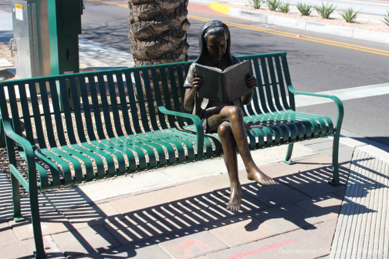 Sculpture of a sitting woman reading a book. She sits on a real park bench.