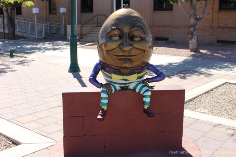 Sculpture of a Humpty Dumpty figure with a big bronze head and colourful clothing sitting on a brick wall