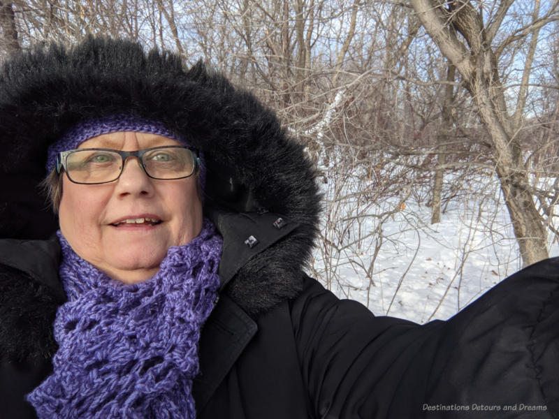 Woman in black parka and purple scarf in front of snow and trees