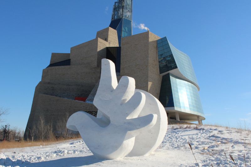 Snow sculpture showing a hand with open fingers in front of the Canadian Museum for Human Rights building