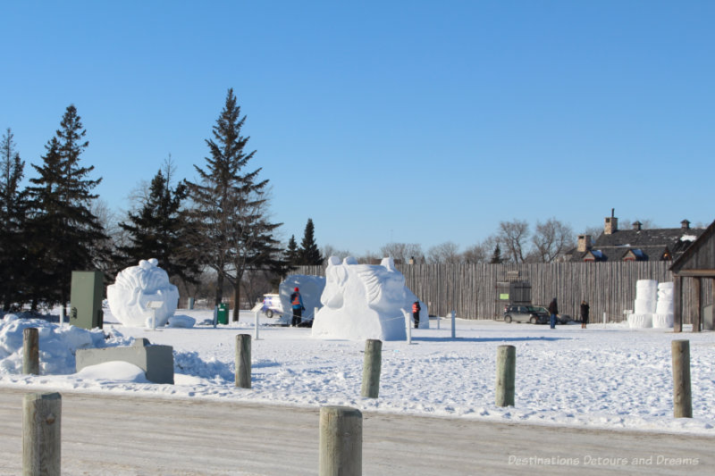 Snow sculptures on the grounds of a park in front of the wooden wall of a fort at the 2021 Festival du Voyageur