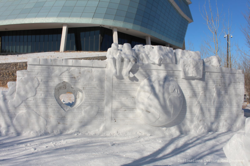 Snow sculpture featuring a wall with a globe and people sketched into it and people sitting atop it