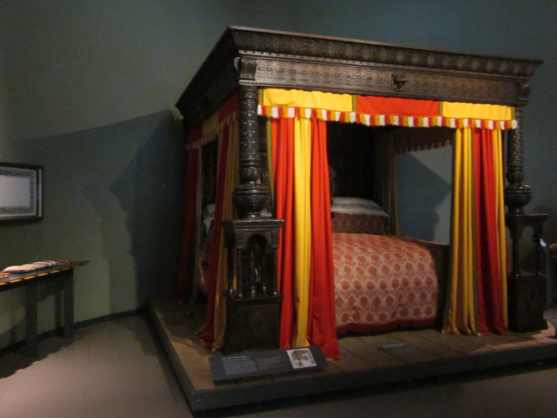 Wooden carved bed of the 1590s with pillars on all four bedposts and red and yellow curtains to enclose bed