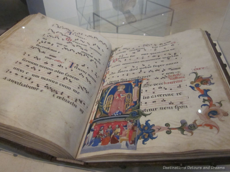 An open choir book from the 14th century showing a decorated page and lines of Latin song words on staffsVicto