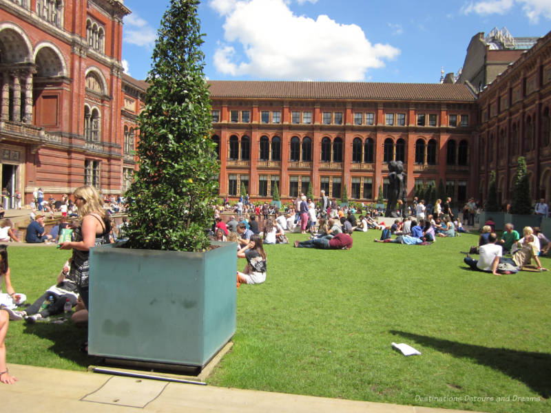 People sitting on the lawn of the V&A courtyard with the beautfiul red stone walls of the museum surrounding them