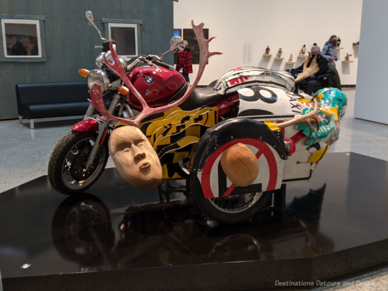 Inuit art piece featuring a motorcycle covered with other pieces such as antlers, a wood sculpted face, and more