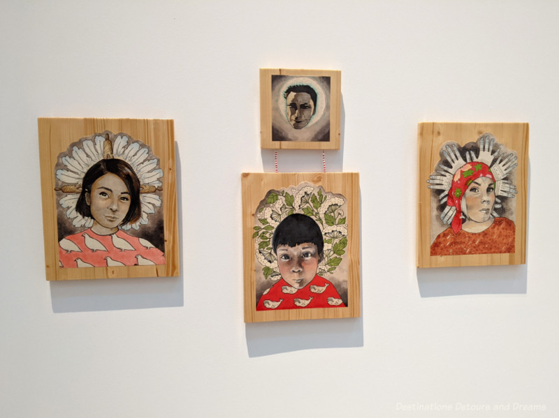 Four wood panel art pieces by Maya Sialuk painted with faces with halos of old Inuit culture items encircling their heads 