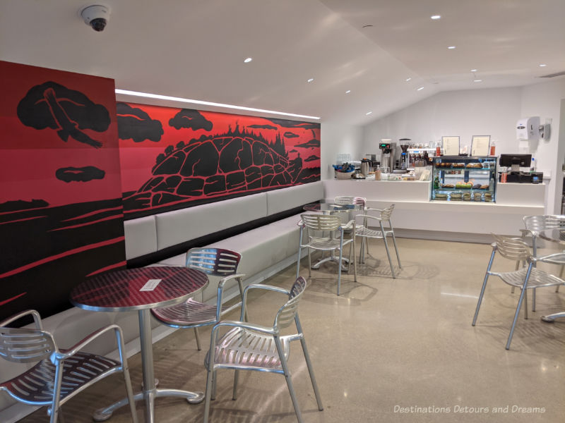 Qaumajuq Café Café at Qaumajuq with food counter, chrome round tables and chairs, and a red and black Inuit art piece along one wallQau