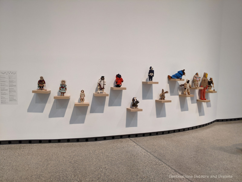 Qaumajuq Inuit Dolls Collection of Inuit dolls displayed on individual wood shelves on the side of large white wall at Qaumajq